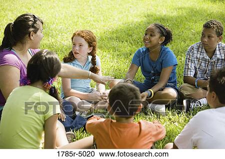 Stock Image - Children and their teacher sitting in a circle on the grass at a park. Fotosearch