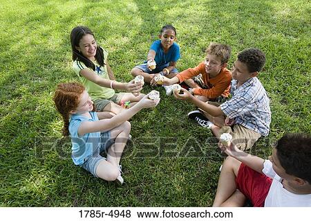 Stock Photo - Children seated in a circle cheering over cupcakes at a park. Fotosearch