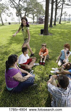 Children sitting in a circle on the grass with their teacher while a girl walks around them View Large Photo Image Picture - Children sitting in a circle on the grass with their teacher while a girl walks around them. Fotosearch