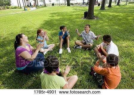 Stock Image - Children sitting in a circle on the grass with their teacher at a park. Fotosearch