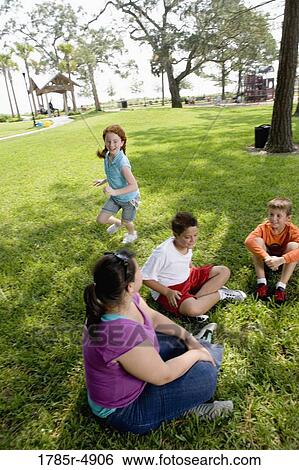 Stock Photograph - Children sitting in a circle on the grass with their teacher while a girl runs around them. Fotosearch