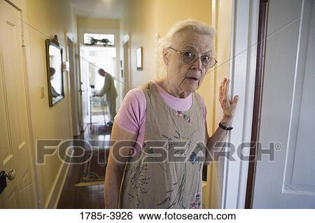Stock Photograph - Elderly woman standing in hallway with feeble senior man in the background. Fotosearch