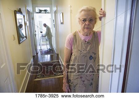 Stock Photography - Elderly woman standing in hallway with feeble senior man in the background. Fotosearch