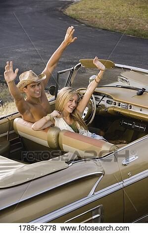 Stock Photo - Elevated view of a young couple waving while sitting in antique convertible car. Fotosearch