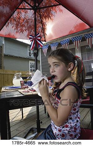 Stock Photography - Girl eating a popsicle at picnic table in backyard patio with 4th of July decorations. Fotosearch