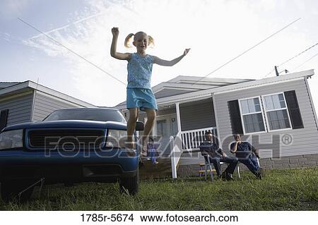 Picture - Girl jumping off bumper of old truck parked in front of trailer home, family in background. Fotosearch