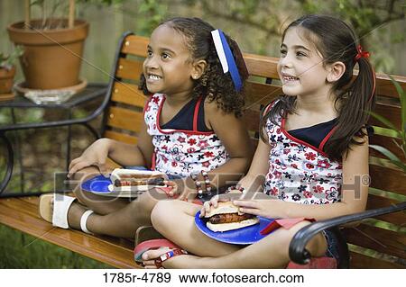 Girls sitting on bench with with a plate of hot dogs on their lap View Large Photo Image Stock Photo - Girls sitting on bench with with a plate of hot dogs on their lap. Fotosearch