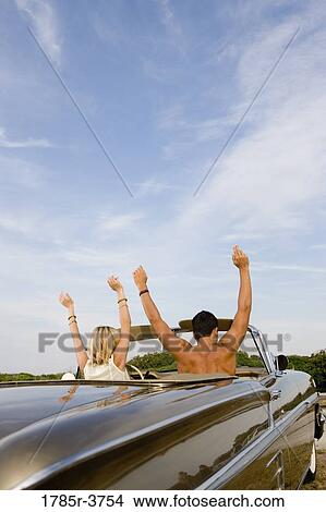 Picture - Happy, cheerful and waving young couple sitting in antique convertible car. Fotosearch