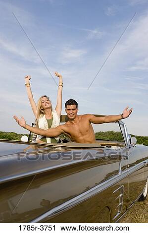 Stock Image - Happy, cheerful and waving young couple sitting in antique convertible car. Fotosearch