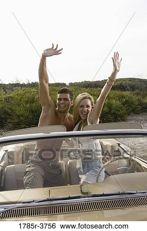 Stock Photograph - Happy, cheerful and waving young couple sitting in antique convertible car. Fotosearch