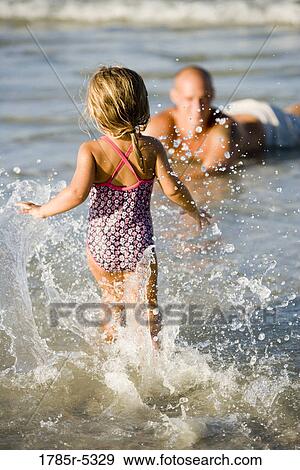 Stock Photo - Little girl splashing in water while father watches at the beach. Fotosearch