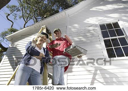 Stock Image - Low angle view of mature couple with ladder and painting tools preparing to paint outside of house. Fotosearch