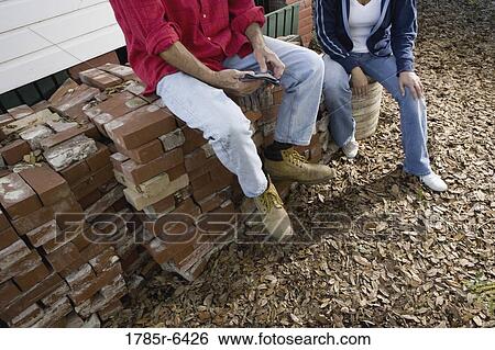 Stock Photograph - Lower portion view of mature couple sitting on stack of bricks outside house. Fotosearch
