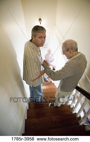 Stock Photo - Man helping elderly man down the stairs while senior woman watches downstairs. Fotosearch