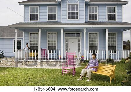 Picture - Mature Asian woman sitting on front lawn of house with husband smoking on porch. Fotosearch