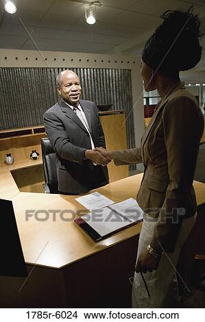 Mature business man standing behind desk shaking hands with younger woman View Large Photo Image Picture - Mature business man standing behind desk shaking hands with younger woman. Fotosearch
