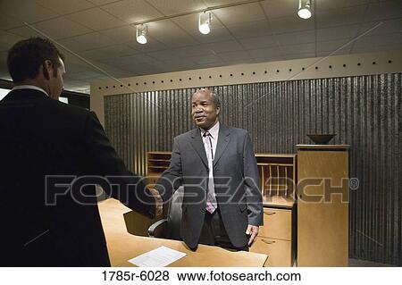 Mature business man standing behind desk shaking hands with younger man View Large Photo Image Stock Photo - Mature business man standing behind desk shaking hands with younger man. Fotosearch