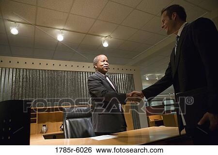 Stock Photograph - Mature business man standing behind desk shaking hands with younger man. Fotosearch