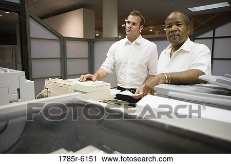 Stock Image - Mature businessman conferring with younger male colleague in an office. Fotosearch