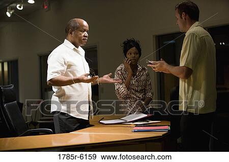 Mature businessman standing behind desk conversing with younger colleagues View Large Photo Image Stock Photo - Mature businessman standing behind desk conversing with younger colleagues. Fotosearch