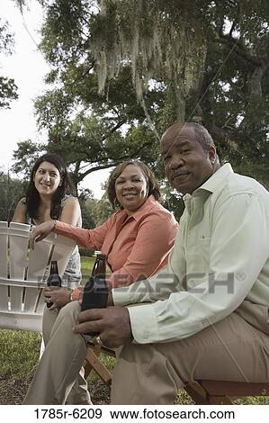 Stock Photo - Mature couple having drinks sitting on lawn chairs next to young woman. Fotosearch