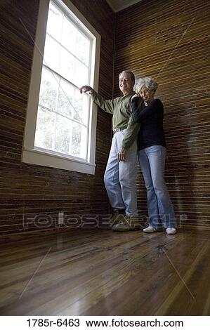 Stock Image - Mature couple in a darkened room leaning against window, gazing out. Fotosearch