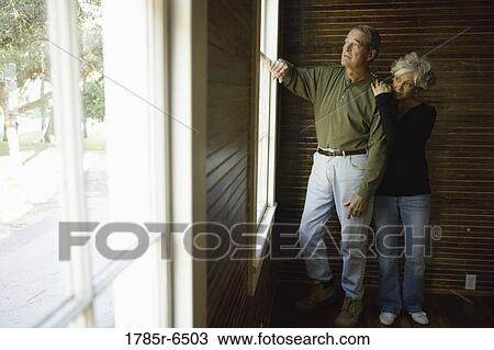 Stock Image - Mature couple in darkened room leaning against window frame looking out. Fotosearch