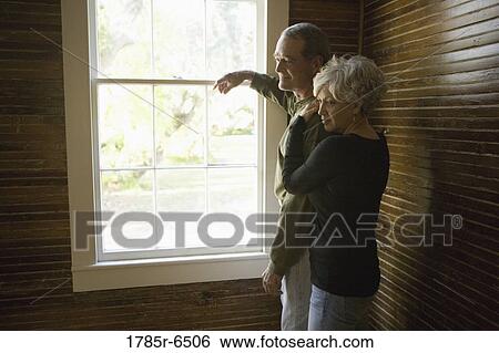 Stock Photograph - Mature couple in darkened room leaning against window frame looking out. Fotosearch