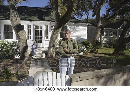 Stock Image - Mature couple in front of house surrounded by tall oak trees. Fotosearch