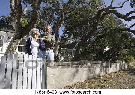 Stock Image - Mature couple in front of house surrounded by tall oak trees. Fotosearch
