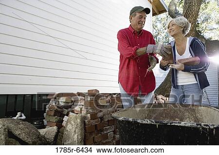 Picture - Mature couple loading bricks on wheelbarrow outside house. Fotosearch