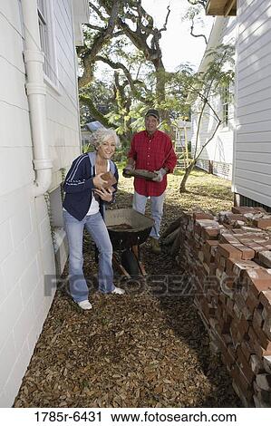 Stock Image - Mature couple loading bricks on wheelbarrow outside house. Fotosearch