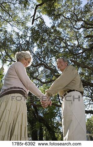 Stock Image - Mature couple standing together under oak trees. Fotosearch
