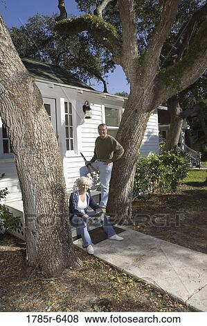 Stock Photo - Mature couple with gardening tools in front of house surrounded by tall oak trees. Fotosearch