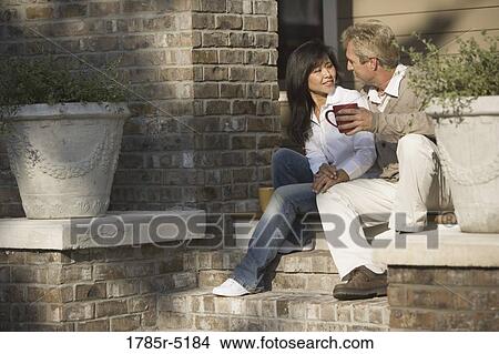 Picture - Mature inter-racial couple holding coffee mugs sitting on front steps of their house. Fotosearch