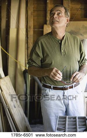 Stock Image - Mature man looking up in tool shed surrounded by lumber and carpentry tools. Fotosearch