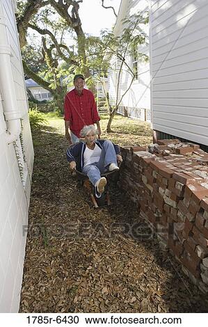 Mature man playfully moving woman sitting in wheelbarrow outside house View Large Photo Image Stock Image - Mature man playfully moving woman sitting in wheelbarrow outside house. Fotosearch
