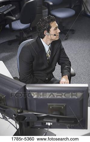 Stock Image - Office worker in a suit with headset sitting at his desk. Fotosearch