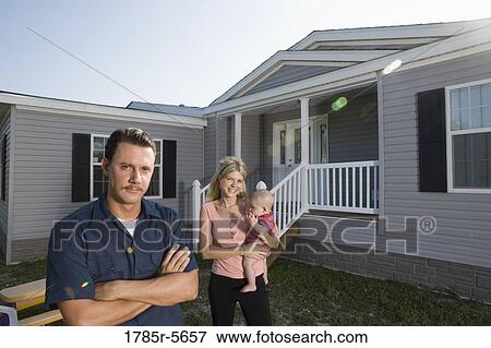 Stock Photo - Portrait of a blue-collar man standing in front of a trailer home with wife and baby. Fotosearch