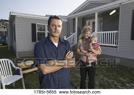 Stock Photography - Portrait of a blue-collar man standing in front of a trailer home with wife and baby. Fotosearch