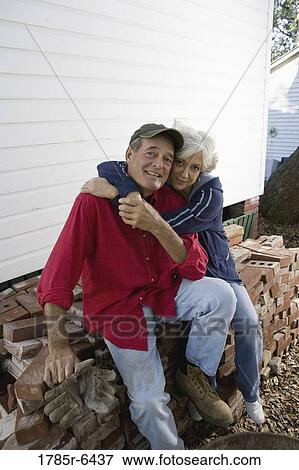 Stock Photo - Portrait of a mature couple embracing while sitting on stack of bricks outside house. Fotosearch