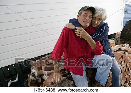 Stock Photo - Portrait of a mature couple embracing while sitting on stack of bricks outside house. Fotosearch