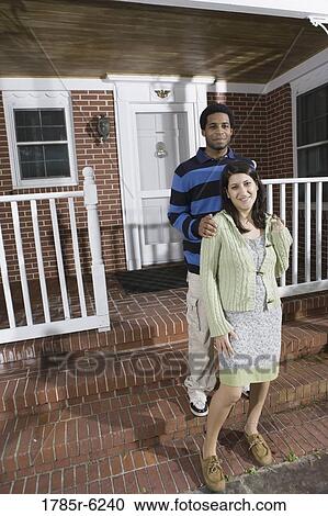 Stock Image - Portrait of a smiling young interracial couple on front steps house. Fotosearch