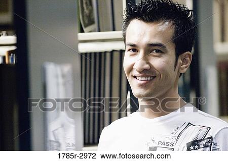 Stock Image - Portrait of a smiling young man amidst library book stacks. Fotosearch