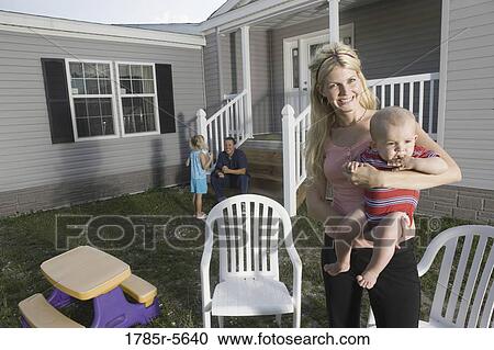 Stock Image - Portrait of a woman holding a baby in front of a trailer home. Fotosearch