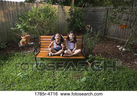 Stock Photograph - Portrait of cheerful girls (6-7) sitting on a bench in a backyard. Fotosearch