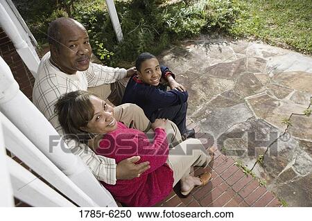 Stock Image - Portrait of mature couple and their son sitting on front steps of house. Fotosearch