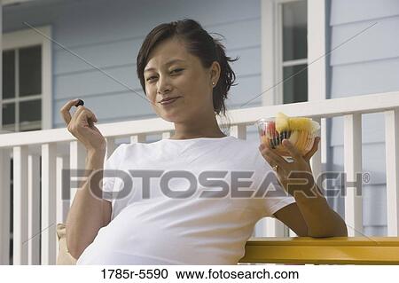 Stock Image - Portrait of pregnant woman holding bowl of fruit in front of house. Fotosearch