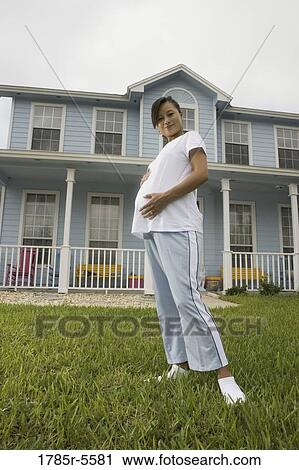 Stock Image - Portrait of pregnant woman standing on lawn in front of house. Fotosearch