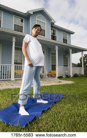 Stock Image - Portrait of pregnant woman standing on yoga mat in front of house. Fotosearch